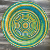 Colorful round hand-braided jute area rug with concentric circles in blue, green, yellow, orange, and white. Eco-friendly, reversible.