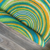 Colorful hand-braided round jute rug featuring a concentric design, partially rolled on a wooden floor. Reversible & eco-friendly.
