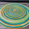 Colorful hand-braided round jute area rug with concentric circles in green, blue, yellow, and natural tones on a wooden floor.