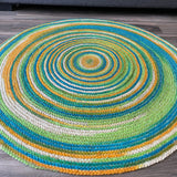 Colorful hand-braided round jute area rug with concentric circles in green, blue, yellow, and natural tones on a wooden floor.