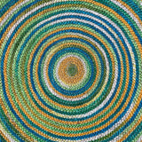 Top-down close-up view of a colorful jute area rug with concentric circles in green, blue, yellow, and natural tones, reversible & eco-friendly.