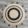 Handwoven round natural jute rug with black concentric circles and alternating natural and black tassels on a gray wood floor.