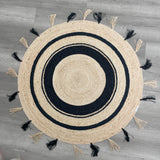 Handwoven round natural jute rug with black concentric circles and alternating natural and black tassels on a gray wood floor.