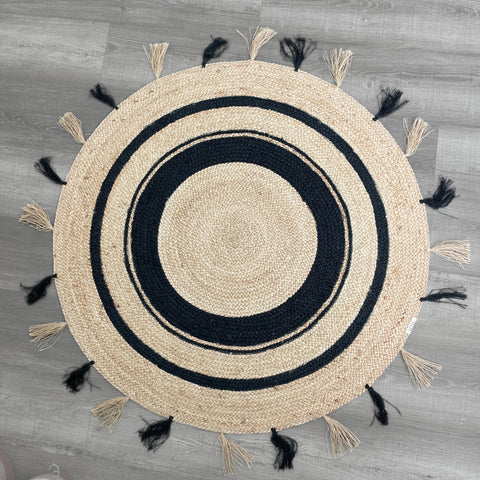 Handwoven round natural jute rug with black concentric circles and alternating natural and black tassels on a gray wood floor.