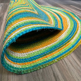 Reversible Round Jute Rug, hand-braided with green, blue, orange, and white stripes, partially rolled on a wooden floor. 